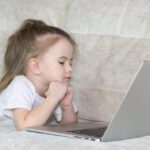 A female Caucasian child with elbows propped on a table whilst looking at a laptop screen