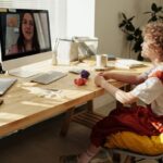 A child looking at a laptop as if interacting with a female adult, presumably a teacher, as if on a video call