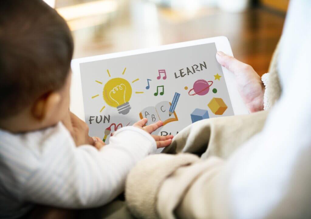 Back view of a parents and a toddler looking at a picture book with drawings of Saturn, a light bulb, etc., with the words FUN and LEARN