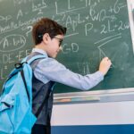 A school-age boy wearing MiyoSmart lenses a backpack and looking confident solving mathematical equations on the blackboard