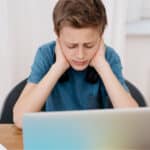 A preteen male child sitting and looking at a laptop, with his hands on the sides of his neck, looking confused or frustrated