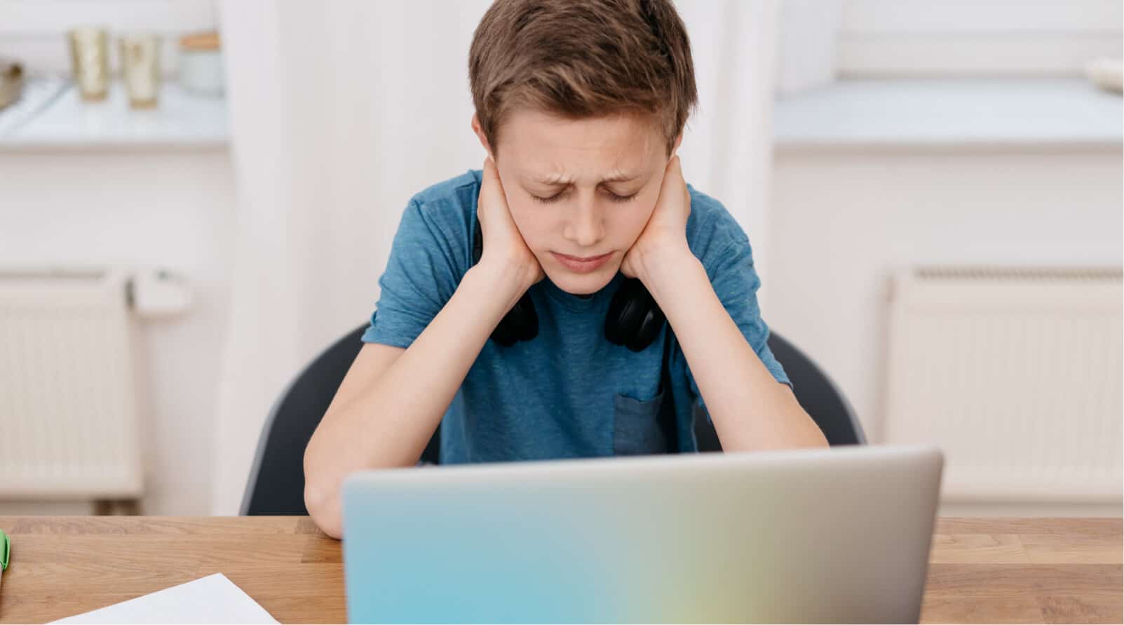 A preteen male child sitting and looking at a laptop, with his hands on the sides of his neck, looking confused or frustrated